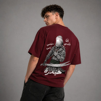 A close-up of a person wearing a burgundy crewneck t-shirt with a white embroidered dagger graphic on the left chest, featuring intricate Arabic calligraphy.