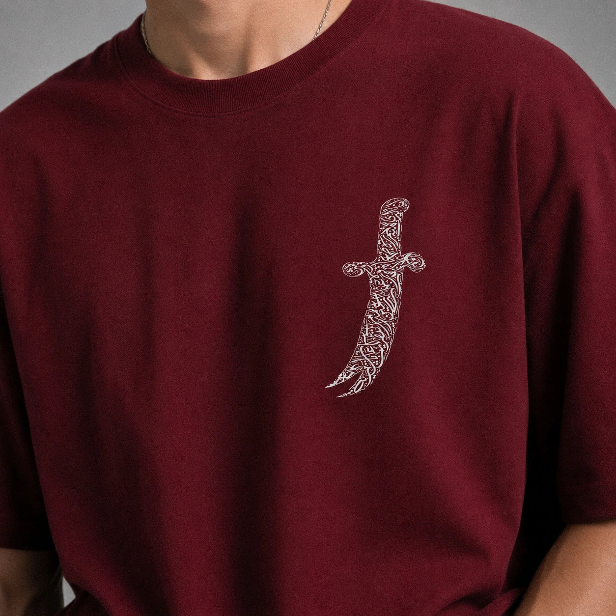 A close-up portrait of a young man with curly brown hair wearing a burgundy crewneck sweatshirt with a silver embroidered sword graphic on the chest, set against a solid gray studio background.