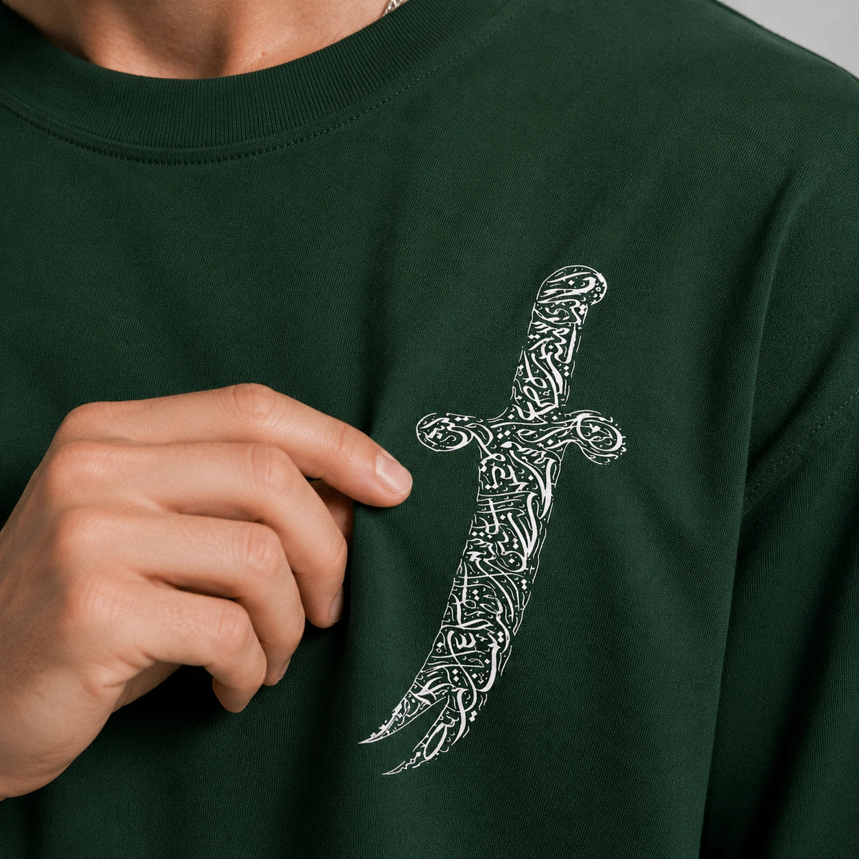 Person wearing a dark green sweatshirt with a white dagger design, pointing to the design with their finger.