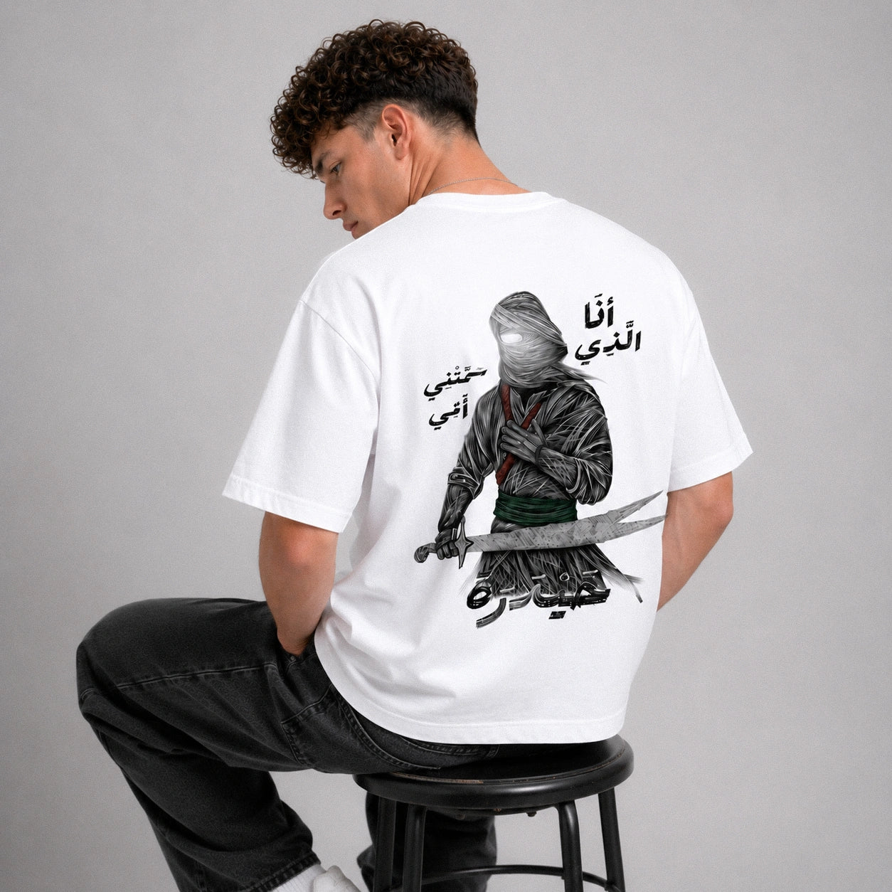 Person wearing a white t-shirt with a graphic design and text on the back, sitting on a stool against a gray background.