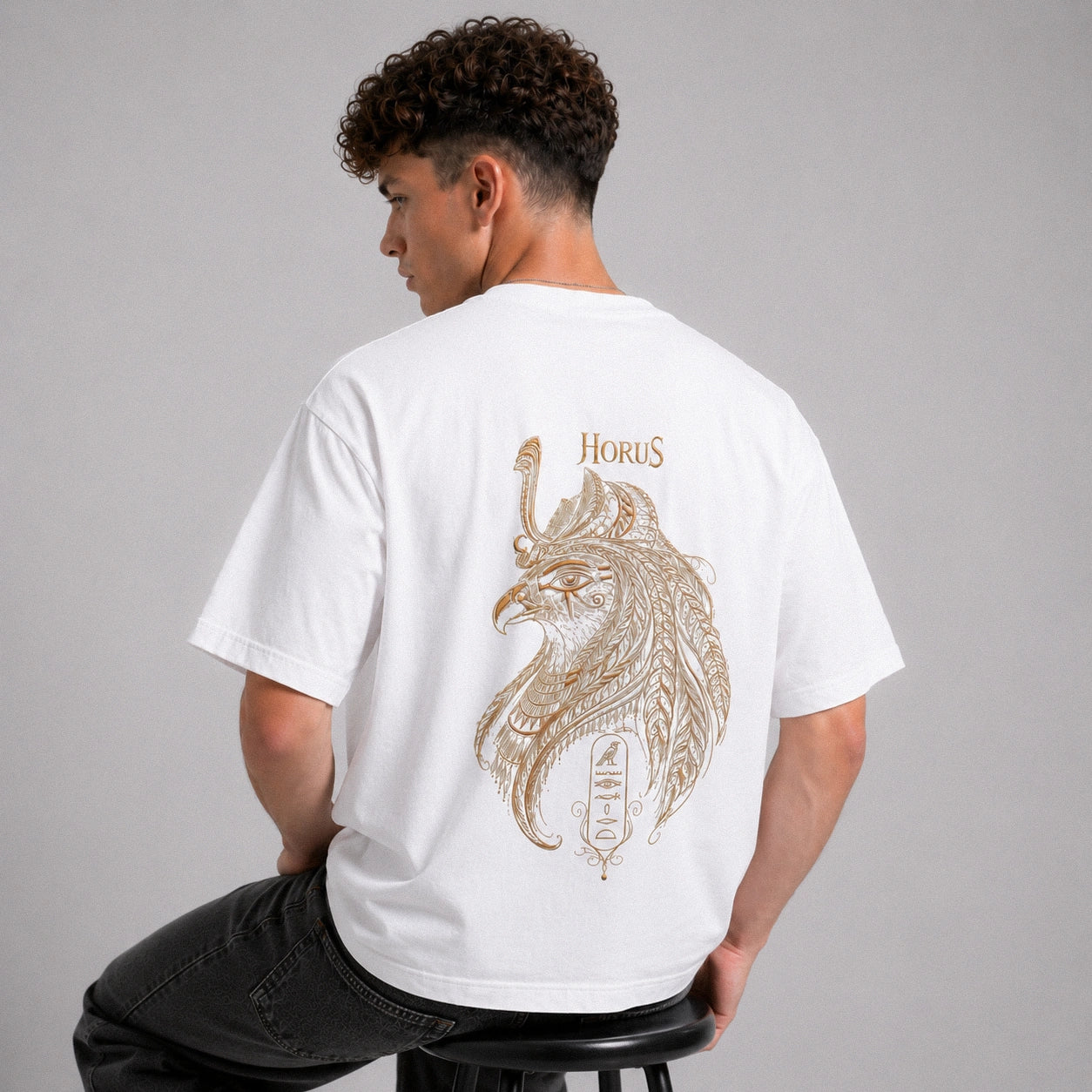 Person wearing a white t-shirt with a Horus design on the back, sitting on a stool against a gray background.