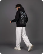 Person wearing a black hoodie with a graphic design on the back, white pants, and white shoes against a gray background