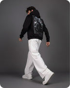 Person wearing a black hoodie with a graphic design on the back, white pants, and white shoes against a gray background