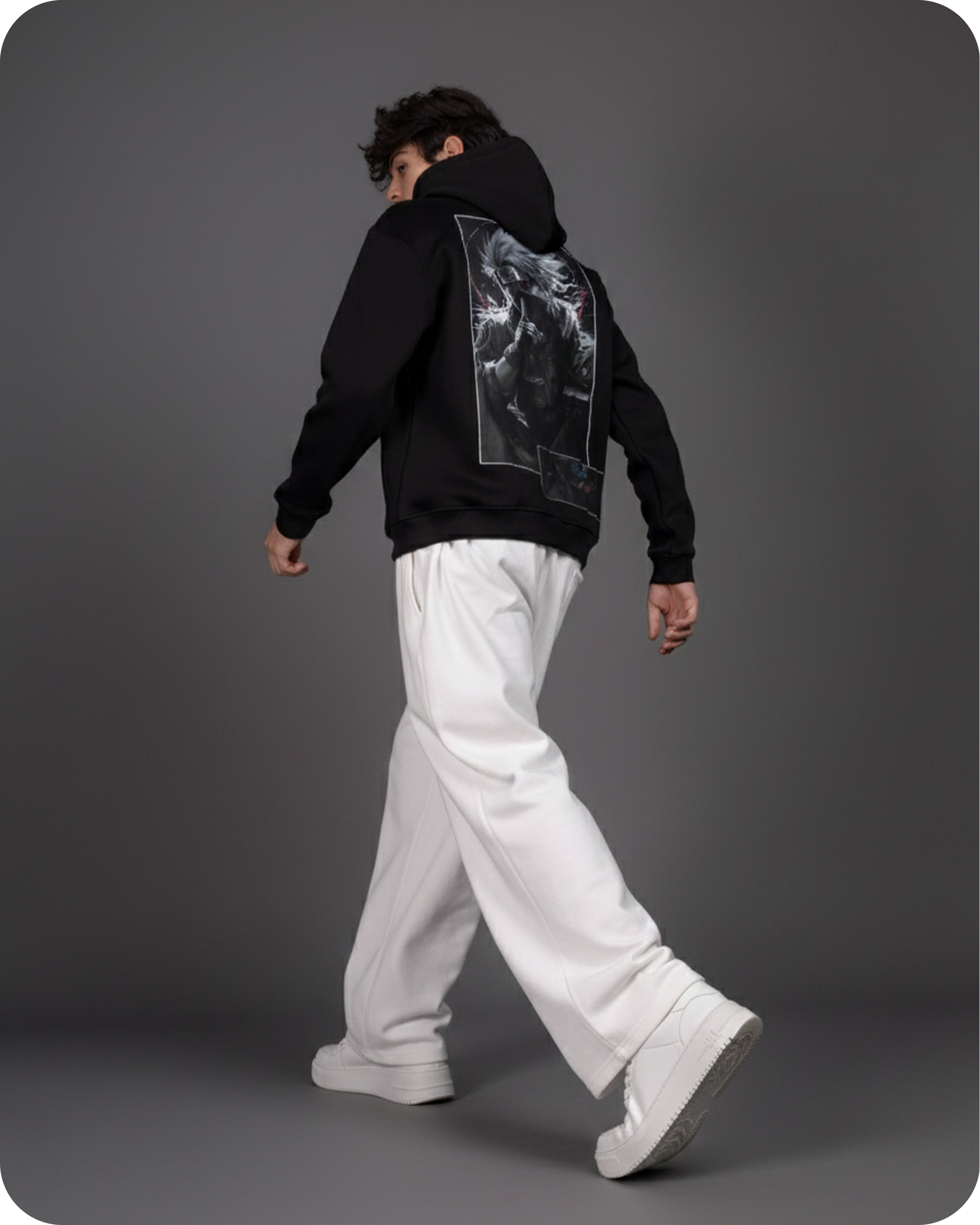 Person wearing a black hoodie with a graphic design on the back, white pants, and white shoes against a gray background