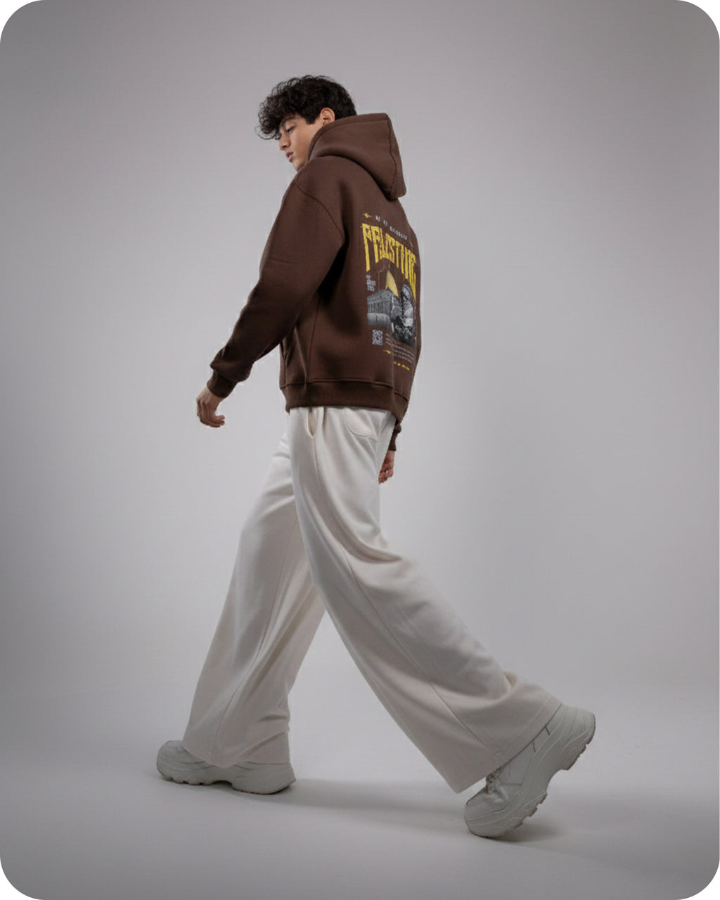 Person wearing a brown hoodie with text and graphics, white pants, and gray shoes on a plain background