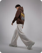 Person wearing a brown hoodie with text and graphics, white pants, and gray shoes on a plain background