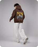 Person wearing a brown hoodie with a graphic design on the back, standing against a gray background