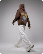 Person wearing a brown hoodie with a graphic design on the back, standing against a gray background