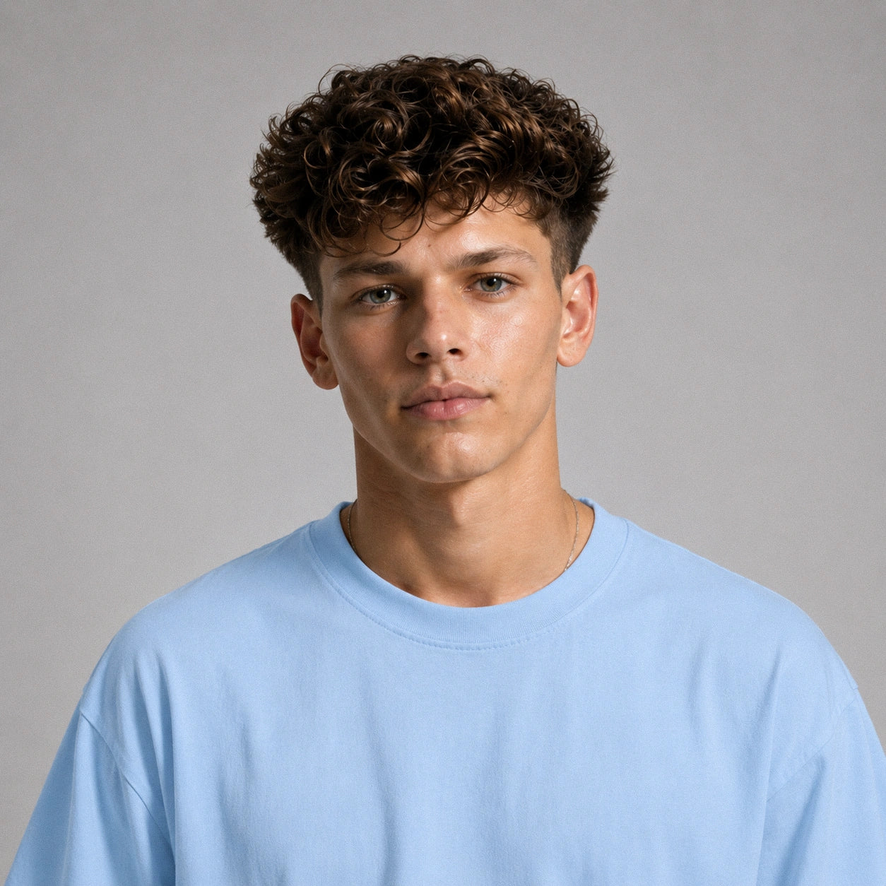 A young man with curly brown hair wearing a light blue crewneck t-shirt, photographed against a neutral gray studio background.