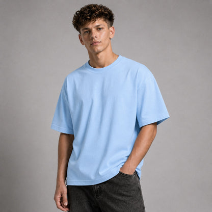 A young man with curly brown hair wearing an oversized light blue crewneck t-shirt and dark gray jeans, standing against a neutral gray studio background.