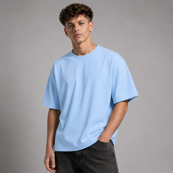 A young man with curly brown hair wearing an oversized light blue crewneck t-shirt and dark gray jeans, standing against a neutral gray studio background.