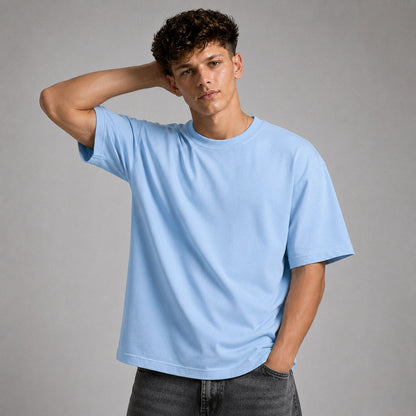 A young man wearing a light blue crewneck t-shirt and dark gray jeans, posing against a solid gray studio background.