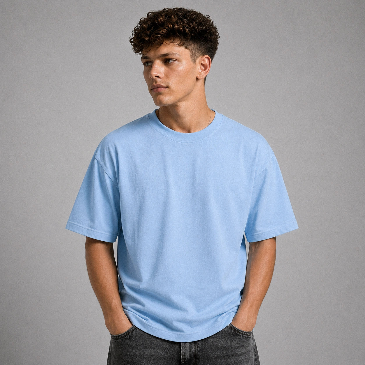 A man wearing a light blue crew-neck t-shirt and dark gray jeans, standing against a solid gray studio background.