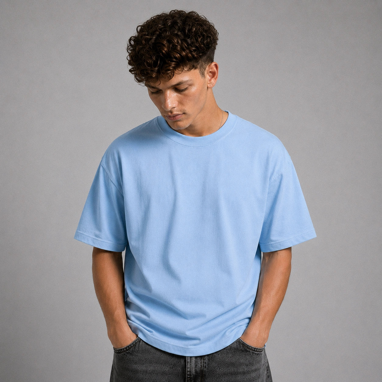 A man wearing a light blue crew-neck t-shirt and dark gray jeans, standing against a solid gray studio background.