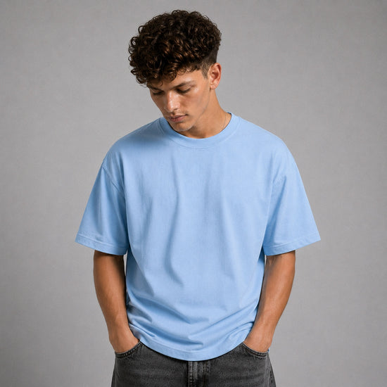 A man wearing a light blue crew-neck t-shirt and dark gray jeans, standing against a solid gray studio background.