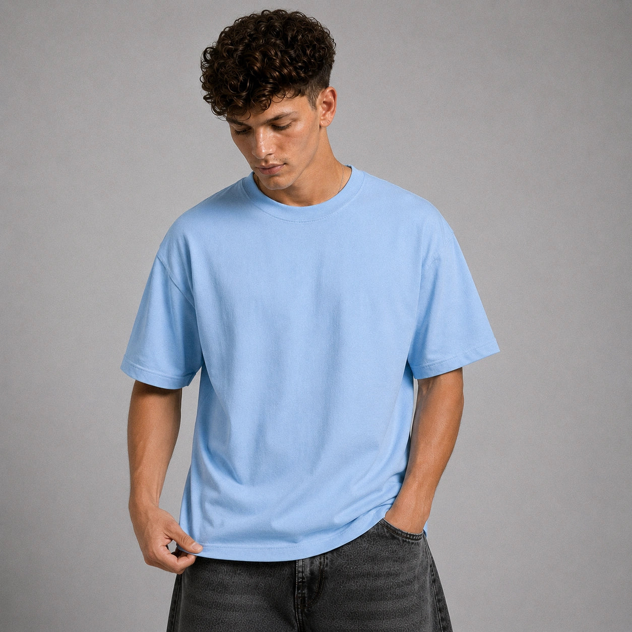 A man wearing a light blue crewneck t-shirt and dark gray jeans, standing against a solid gray studio background.