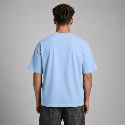 A man viewed from behind wearing a light blue crewneck t-shirt and dark gray jeans, standing against a neutral gray studio background.