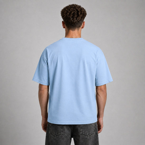 A man viewed from behind wearing a light blue crewneck t-shirt and dark gray jeans, standing against a neutral gray studio background.