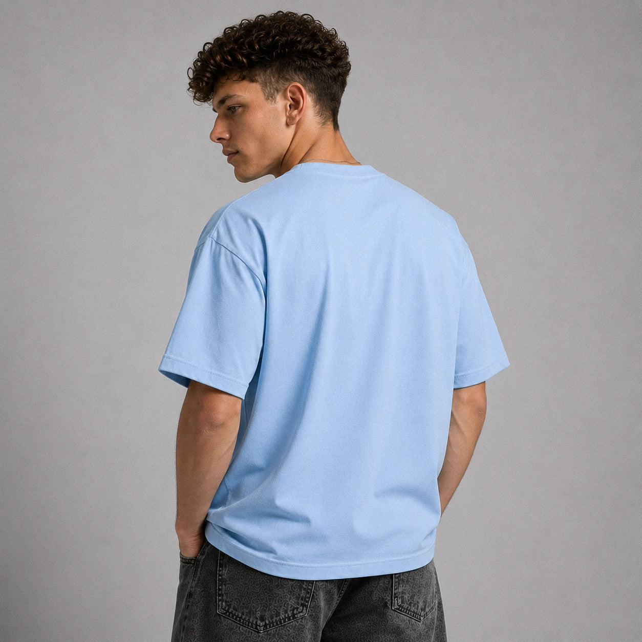 A man with curly dark hair is shown from the back wearing a light blue crewneck t-shirt and dark gray jeans, standing against a solid gray studio background.