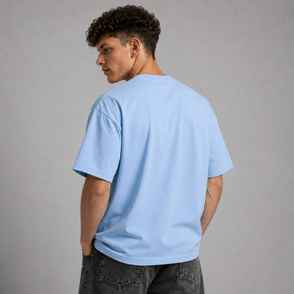 A man with curly dark hair is shown from the back wearing a light blue crewneck t-shirt and dark gray jeans, standing against a solid gray studio background.
