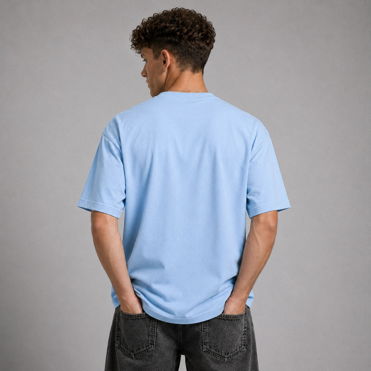 A man viewed from behind wearing a light blue crewneck t-shirt and dark gray jeans, standing against a solid gray studio background.