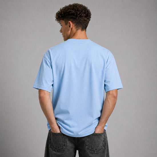 A man viewed from behind wearing a light blue crewneck t-shirt and dark gray jeans, standing against a solid gray studio background.