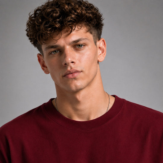 A close-up portrait of a young man with curly brown hair wearing a crewneck sweatshirt in deep burgundy against a neutral gray background.