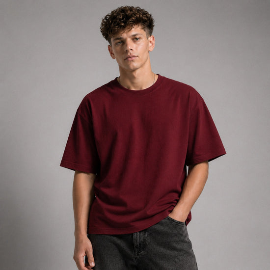 A young man with curly brown hair wearing an oversized, short-sleeve crewneck t-shirt in deep burgundy, paired with dark gray jeans, standing against a solid gray studio background.