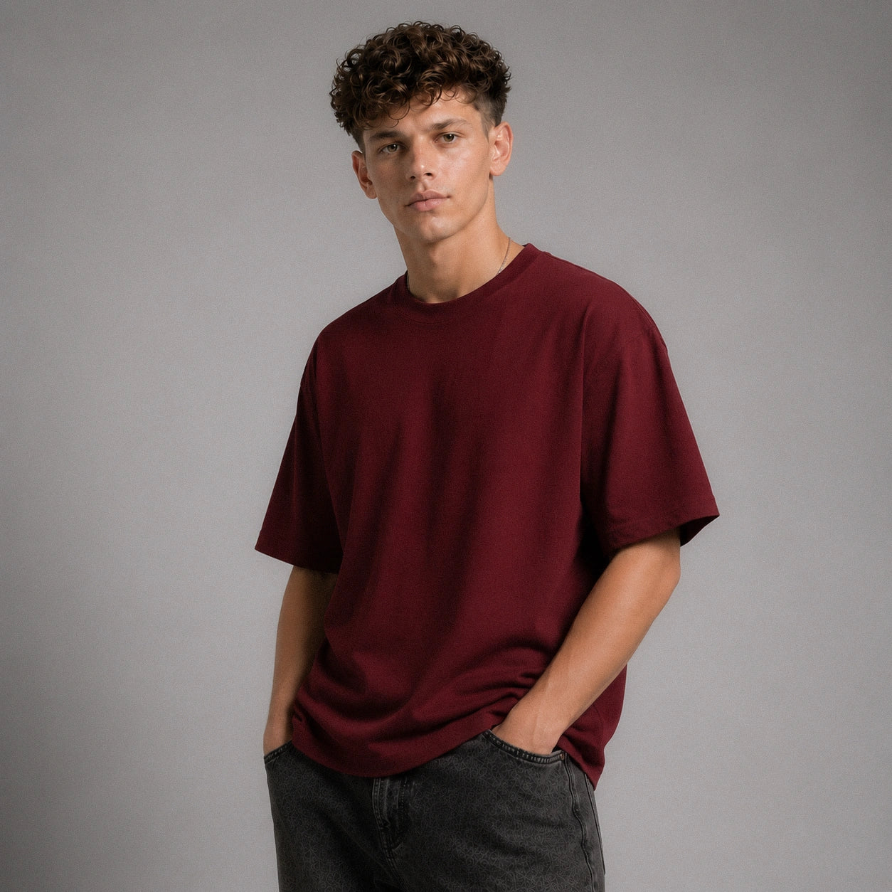 A young man with curly brown hair wearing an oversized maroon crewneck t-shirt and dark gray jeans, standing against a solid gray studio background.