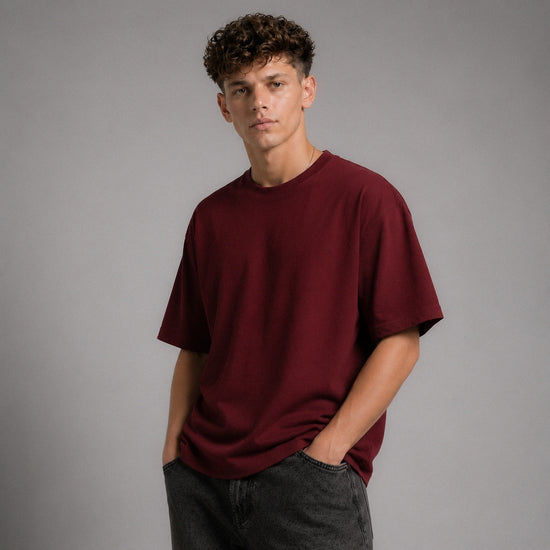 A young man with curly brown hair wearing an oversized maroon crewneck t-shirt and dark gray jeans, standing against a solid gray studio background.