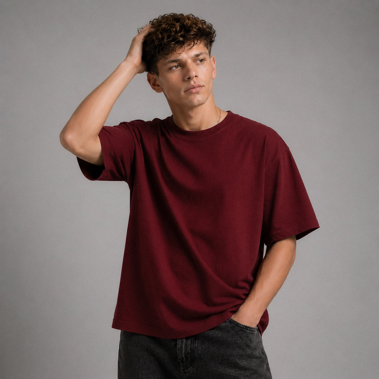 A man wearing an oversized maroon short-sleeve t-shirt and dark gray jeans, posing against a solid gray studio background.