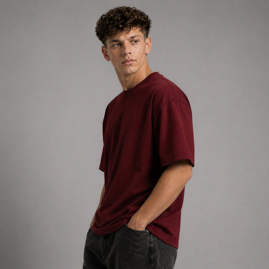 A young man with curly brown hair wearing an oversized, short-sleeve crewneck t-shirt in deep burgundy, paired with dark gray jeans, standing against a solid gray studio background.