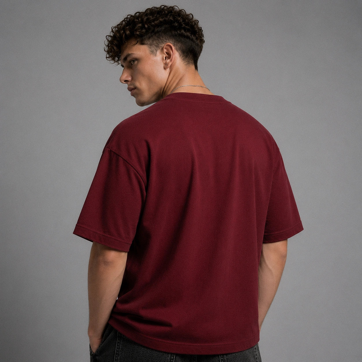 A man with curly dark hair is shown from the back wearing an oversized short-sleeve crewneck t-shirt in a deep burgundy or maroon color, paired with dark jeans, against a solid gray studio background.