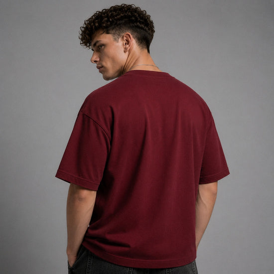 A man with curly dark hair is shown from the back wearing an oversized short-sleeve crewneck t-shirt in a deep burgundy or maroon color, paired with dark jeans, against a solid gray studio background.