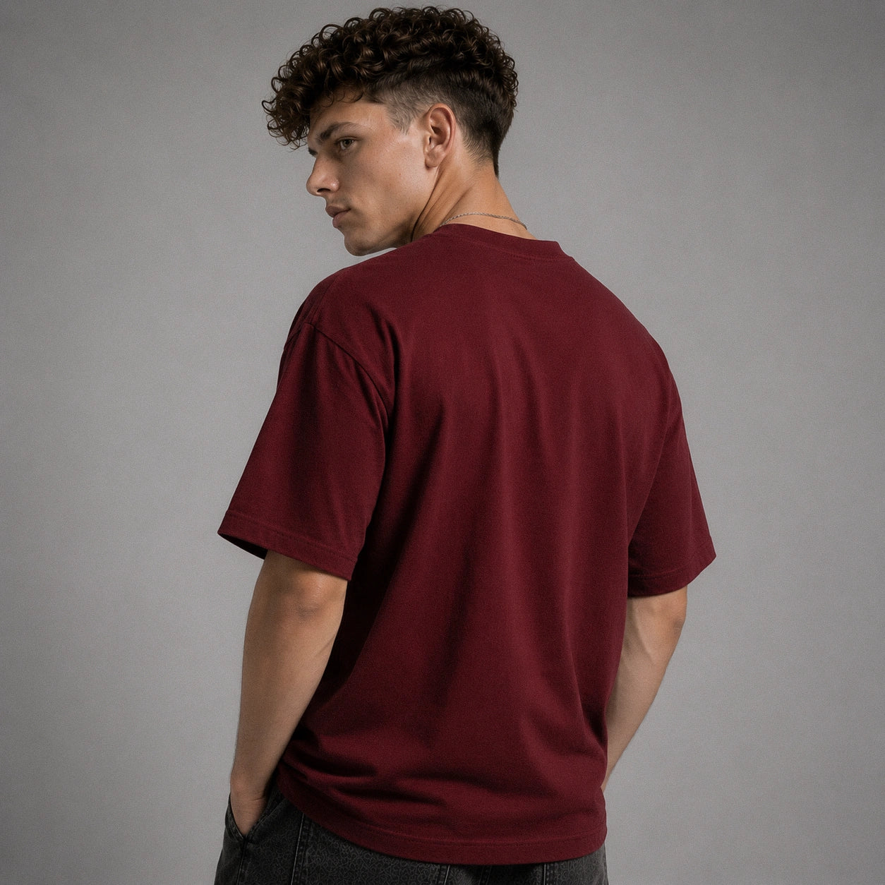 A man with curly dark hair is shown from the back wearing a plain maroon short-sleeve t-shirt and dark jeans, standing against a solid gray studio background.