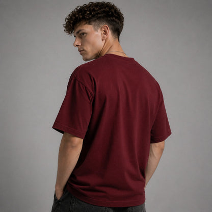 A man with curly dark hair is shown from the back wearing a plain maroon short-sleeve t-shirt and dark jeans, standing against a solid gray studio background.