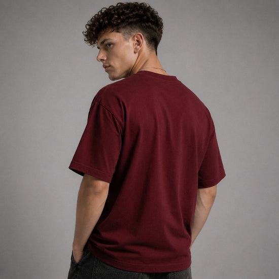 A man with curly dark hair is shown from the back wearing a plain maroon short-sleeve t-shirt and dark jeans, standing against a solid gray studio background.