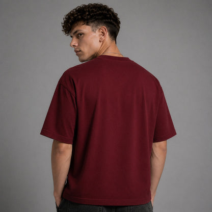 A man wearing an oversized short-sleeve crewneck t-shirt in maroon, shown from the back against a solid gray studio background.