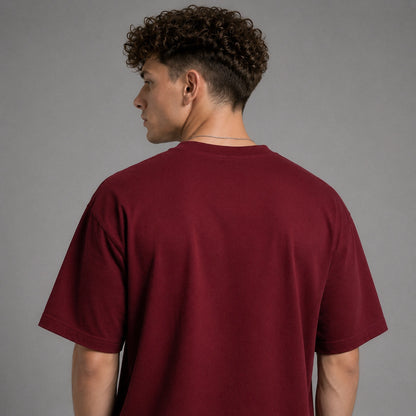 A man with curly brown hair is shown from the back wearing a solid maroon short-sleeve crewneck t-shirt against a plain gray studio background.