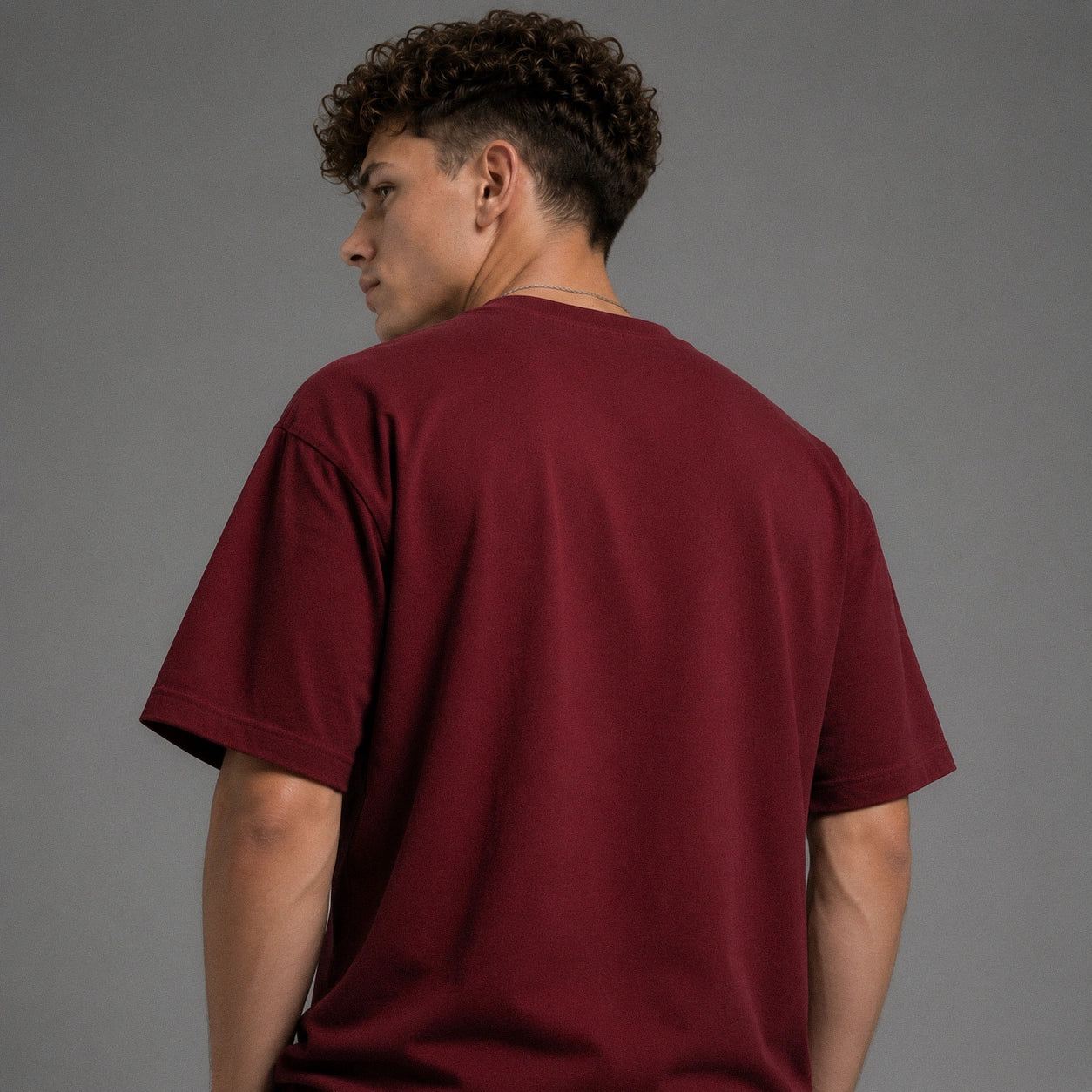A man with curly dark hair is shown from the back wearing a short-sleeve crewneck t-shirt in a deep burgundy or maroon color, set against a plain gray studio background.