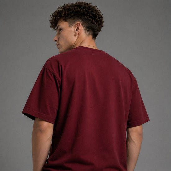 A man with curly dark hair is shown from the back wearing a short-sleeve crewneck t-shirt in a deep burgundy or maroon color, set against a plain gray studio background.