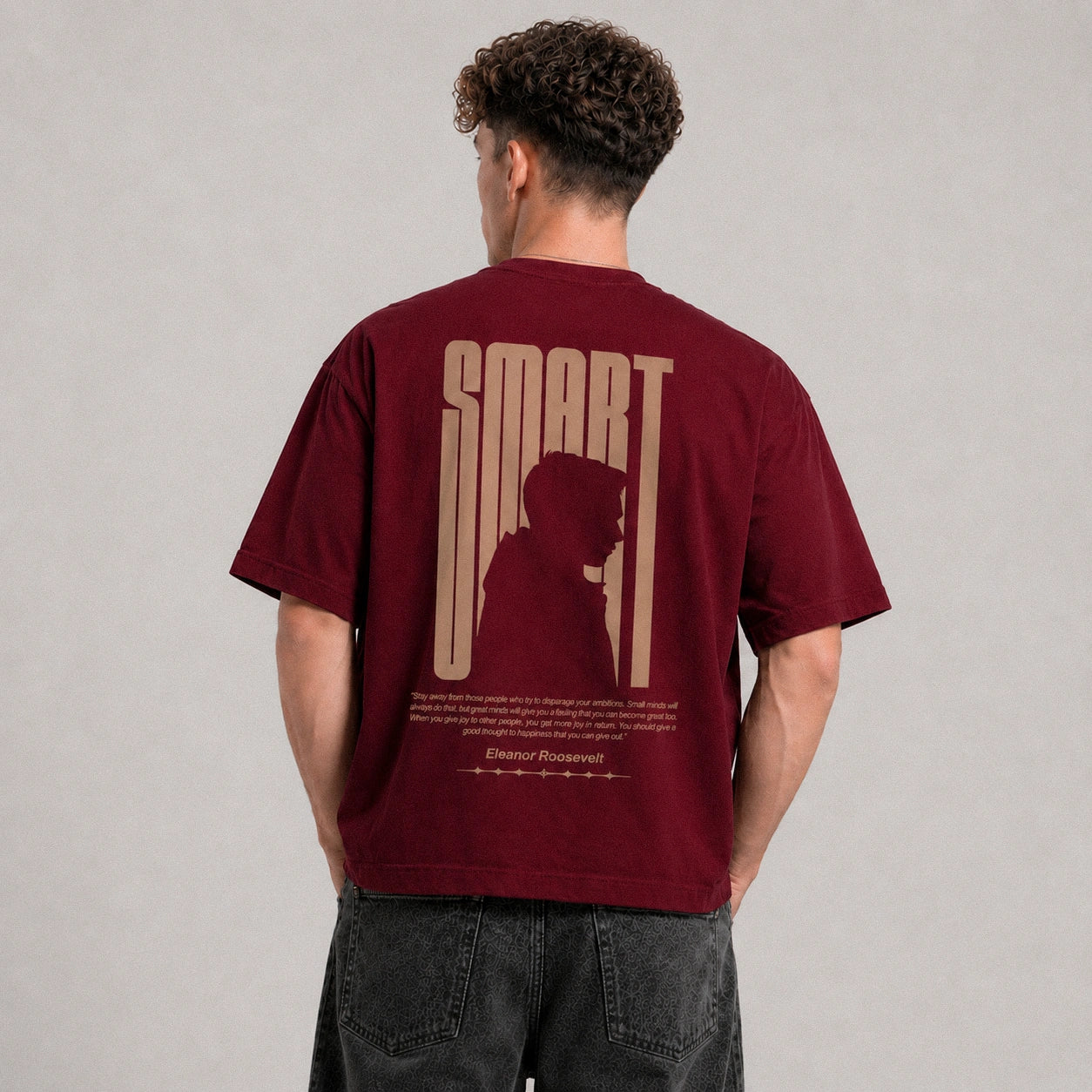 A man viewed from behind wearing a maroon short-sleeve t-shirt with a large beige graphic on the back that reads 'SMART' and features a silhouette of a person. He is also wearing dark gray jeans against a neutral gray studio background.