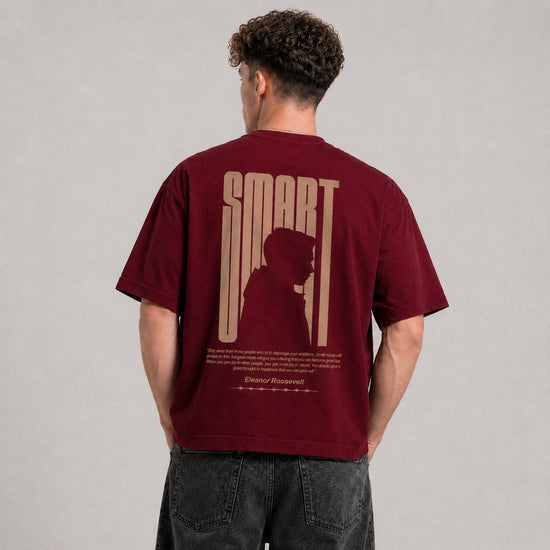 A man viewed from behind wearing a maroon short-sleeve t-shirt with a large beige graphic on the back that reads 'SMART' and features a silhouette of a person. He is also wearing dark gray jeans against a neutral gray studio background.