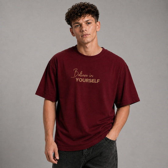 A young man with curly brown hair wearing an oversized maroon t-shirt with 'Believe in YOURSELF' printed in gold on the chest, paired with dark gray jeans, against a solid gray studio background.