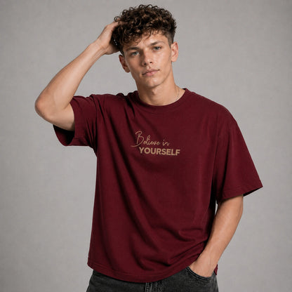 A young man with curly brown hair wearing an oversized maroon t-shirt with the words 'Believe in YOURSELF' printed in gold on the chest, posed against a neutral gray background.
