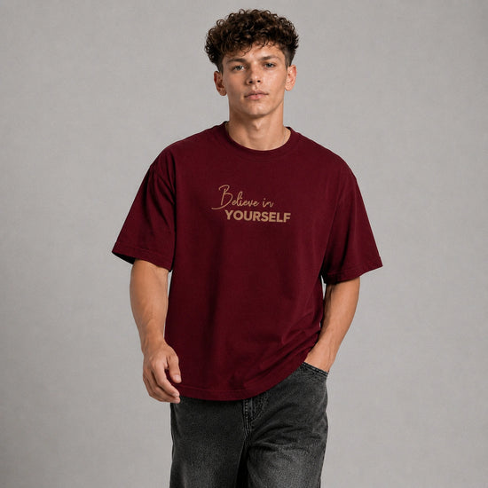 A young man with curly brown hair wearing an oversized maroon t-shirt with 'Believe in YOURSELF' printed in gold on the chest, paired with dark gray jeans, against a solid gray studio background.