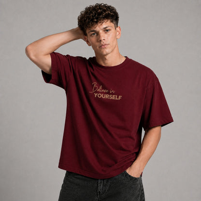 A young man with curly brown hair wearing a maroon crewneck t-shirt with 'Believe in YOURSELF' printed in gold on the chest, posed against a neutral gray background.