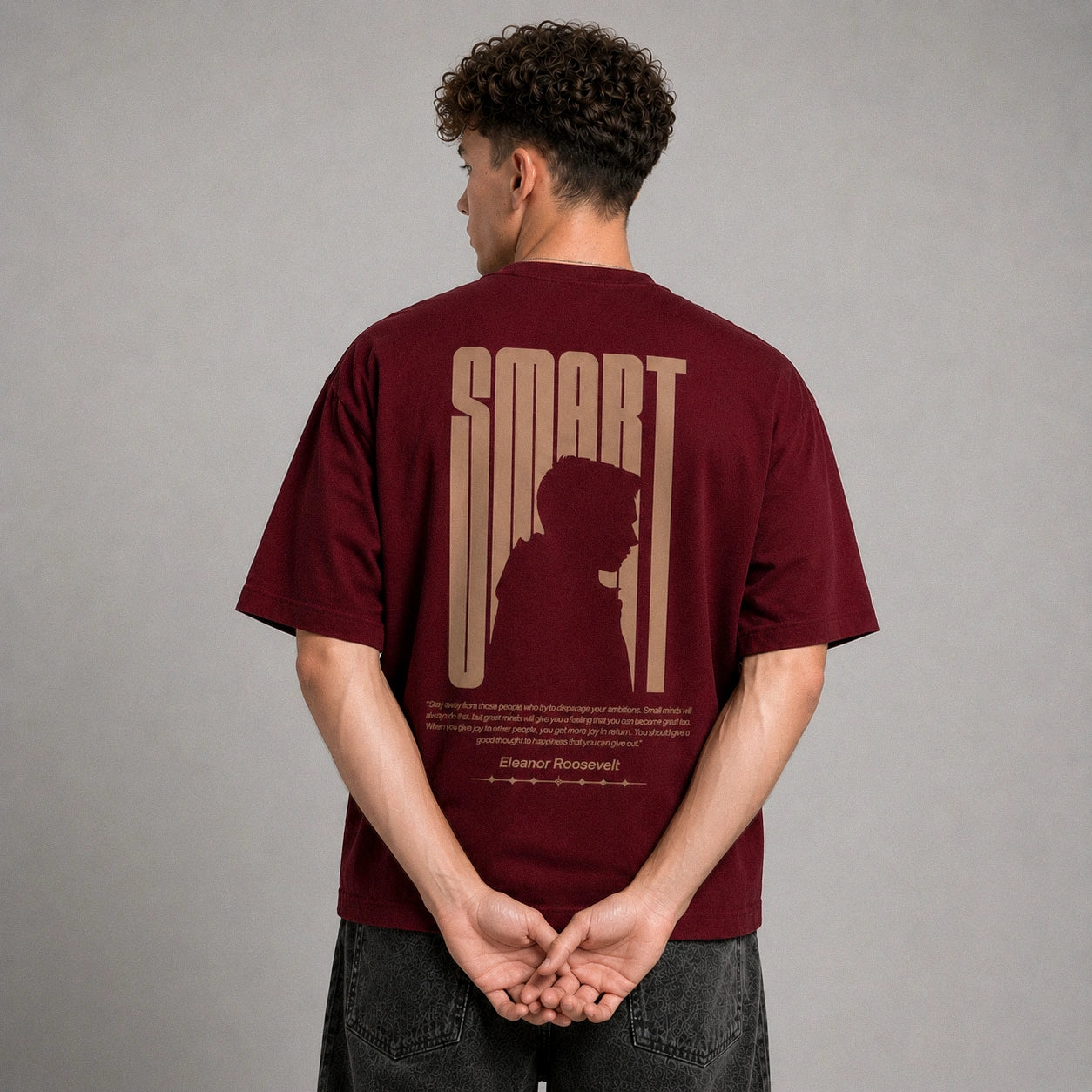 A man viewed from behind wearing a maroon t-shirt with a beige graphic print on the back that reads 'SMART' and features a silhouette of a person. The background is a solid gray studio backdrop.