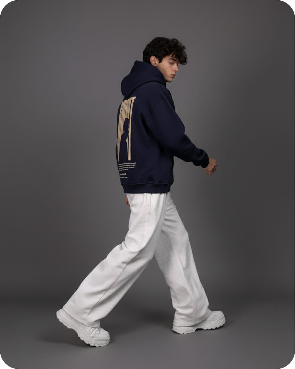 Person wearing a navy hoodie with a graphic design on the back, white pants, and white shoes against a gray background