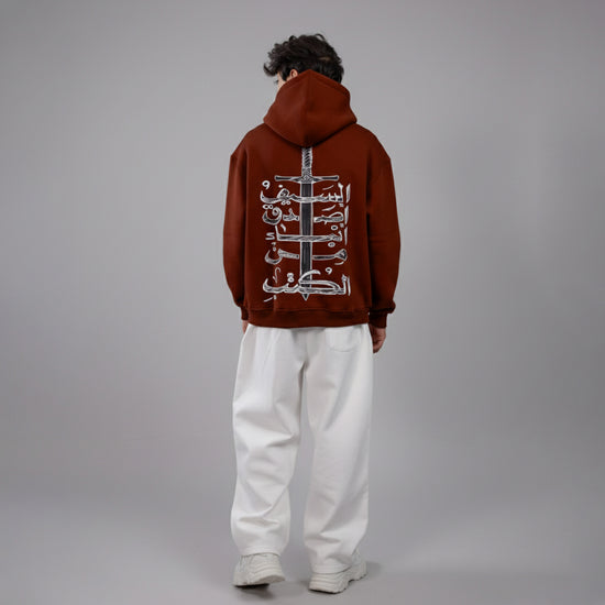 Person wearing a brown hoodie with Arabic text on the back against a gray background